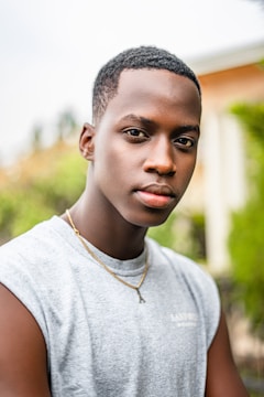 A young person with short hair wearing a light gray sleeveless shirt and a gold necklace is standing outdoors. The background is slightly blurred, showing greenery and part of a building.