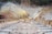 A construction site with a tunnel being built into a hillside. There are wooden beams and concrete structures in the foreground. Dust or smoke is emerging from the rough circular tunnel entrance, with some trees and rocky terrain in the background.