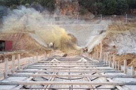A construction site with a tunnel being built into a hillside. There are wooden beams and concrete structures in the foreground. Dust or smoke is emerging from the rough circular tunnel entrance, with some trees and rocky terrain in the background.