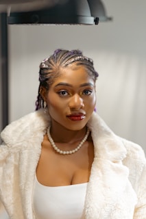 A person with braided hair adorned with beads is wearing a white fur-like coat and a pearl necklace. The background is softly blurred, adding focus to the subject's confident expression and bold makeup, including red lipstick and highlighted eyes.