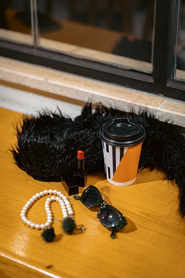 A wooden surface hosts a collection of fashionable accessories, including a black faux fur item, a take-out coffee cup with orange and black geometric patterns, a red lipstick, a string of white pearls, and dark sunglasses. The setting is elegant and organized, suggesting a theme of sophistication.