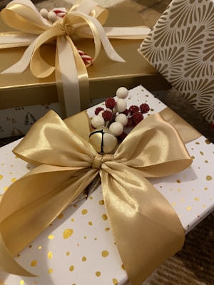 A close-up view of elegantly wrapped gifts featuring large gold satin bows and festive decorations such as a small sprig of red and white berries. The boxes have a luxurious appearance with one adorned in gold and the other with a white and gold polka dot pattern. Another box with a patterned design is partially visible.
