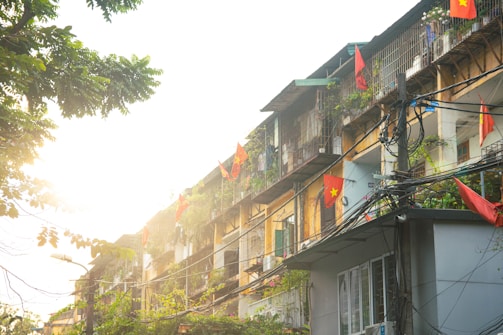 Apartment buildings adorned with numerous flags, likely Vietnamese flags, are seen under bright sunlight. The structures have a mix of metal bars and plants on their balconies. Power lines are prominently visible, adding to the urban setting, and the scene is partially framed by tree branches.