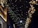 Strings of twinkling warm white fairy lights are hanging in cascading patterns against a dark background, creating a festive and enchanting atmosphere. The soft glow of the lights illuminates part of the building facade they are attached to.