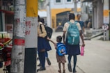 Two schoolgirls are walking down a street, one holding a younger girl's hand. The older girl carries a turquoise backpack and a pink water bottle, while the younger child has a blue backpack with a cartoon design. They are near utility poles with some text and posters. The background shows a building with yellow and white walls, and other people walking in the distance.