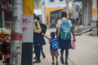 Two schoolgirls are walking down a street, one holding a younger girl's hand. The older girl carries a turquoise backpack and a pink water bottle, while the younger child has a blue backpack with a cartoon design. They are near utility poles with some text and posters. The background shows a building with yellow and white walls, and other people walking in the distance.