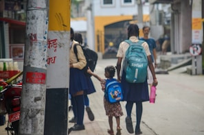 Two schoolgirls are walking down a street, one holding a younger girl's hand. The older girl carries a turquoise backpack and a pink water bottle, while the younger child has a blue backpack with a cartoon design. They are near utility poles with some text and posters. The background shows a building with yellow and white walls, and other people walking in the distance.