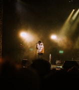 A performer wearing a white puffer jacket and hat stands on stage with a microphone. The stage is dimly lit with warm spotlights creating an atmospheric ambiance. Smoke or mist enhances the dramatic effect, with a few shadowy figures in the background, likely crew members or audience.