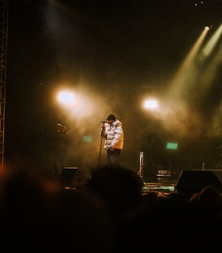 A performer wearing a white puffer jacket and hat stands on stage with a microphone. The stage is dimly lit with warm spotlights creating an atmospheric ambiance. Smoke or mist enhances the dramatic effect, with a few shadowy figures in the background, likely crew members or audience.