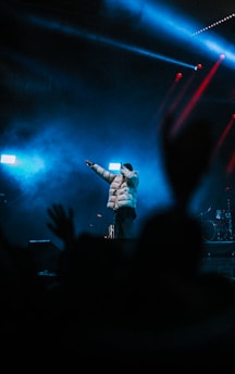 A performer stands on a dimly lit stage, wearing a puffy jacket and a hat. Blue lights and fog create a dramatic atmosphere, while red spotlights add dynamic lighting. Silhouetted hands from the audience are visible in the foreground, conveying a sense of excitement and engagement.