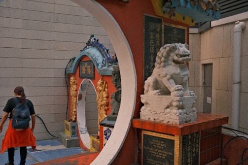A stone guardian lion sits prominently on a pedestal, surrounded by intricately decorated archways featuring bright colors and detailed carvings. A person walks by, wearing a backpack and a red jacket tied around the waist. The setting appears to be a traditional or cultural site.