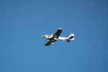 A small, light aircraft with fixed wings and a propeller is flying against a clear blue sky. The aircraft is white with blue accents and features a registration number on its body.