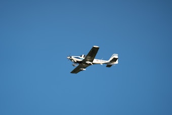 A small, light aircraft with fixed wings and a propeller is flying against a clear blue sky. The aircraft is white with blue accents and features a registration number on its body.