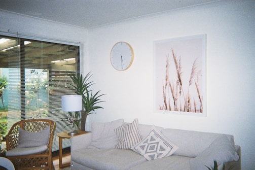 A cozy living room featuring a beige sofa adorned with patterned pillows. Above the sofa, a minimalist clock and a framed print of tall grass add decor to the white wall. A wicker chair and a small wooden table with a lamp are situated nearby, and a tall indoor plant adds greenery. Large windows with blinds open to a view of an outdoor garden.