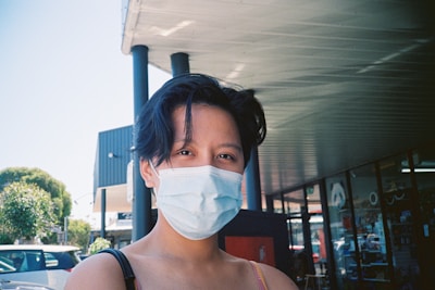 A person with short dark hair is wearing a light blue surgical mask. They are standing outside a building with a shaded awning. In the background, there are several parked cars and greenery, and part of a storefront with glass windows is visible.