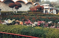 A beautifully landscaped garden featuring a variety of colorful flowers arranged in neat rows. The garden is surrounded by neatly trimmed hedges and ornamental trees, with a stone wall and a structure visible in the background.