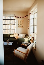 A cozy living room with a green sectional sofa adorned with pillows and a throw blanket. A wooden coffee table holds a few items, including a carafe and coasters. There's a comfortable armchair nearby, and a window ledge with potted plants and framed photos. The walls feature decorative prints and a garland of red paper stars hangs above.