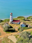 A coastal scene featuring a white and red striped lighthouse situated near a calm body of turquoise water. The lighthouse is surrounded by small buildings with red roofs and thatched coverings, enclosed within a wooden fence. The area is lush with green grass and trees, and a dirt path leads up to the complex, suggesting a serene and isolated setting.