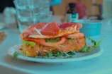 A sandwich on a plate features slices of tomato, greens, and cured meat layered on a seasoned bread roll. The sandwich is placed on a white plate garnished with fresh herbs. In the background, there are a water glass and a blurred container or bottle that appear to be on a table or countertop.