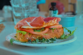A sandwich on a plate features slices of tomato, greens, and cured meat layered on a seasoned bread roll. The sandwich is placed on a white plate garnished with fresh herbs. In the background, there are a water glass and a blurred container or bottle that appear to be on a table or countertop.