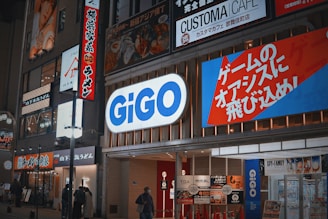 A busy urban street scene at night showcases various illuminated storefronts, with the prominent 'GiGO' sign lit up in bright blue and white. Surrounding it are other signs in Japanese, advertising different businesses including a ramen shop and a café. The street is dimly lit but bustling with activity, featuring people walking and gathering near the entrances.