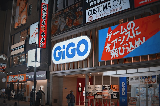 A busy urban street scene at night showcases various illuminated storefronts, with the prominent 'GiGO' sign lit up in bright blue and white. Surrounding it are other signs in Japanese, advertising different businesses including a ramen shop and a café. The street is dimly lit but bustling with activity, featuring people walking and gathering near the entrances.