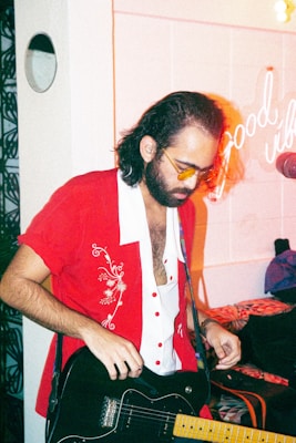 A person wearing a red and white embroidered shirt is playing an electric guitar. They have long hair, a full beard, and are wearing tinted glasses. In the background, neon text reading 'Good vibes' is visible on the wall, and a microphone is positioned nearby.