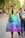 People are walking on a tree-lined street, one person wearing a rainbow flag cape with the word 'Equal' written on it. Others in the background are also casually dressed, potentially attending a public event or parade. The scene conveys an atmosphere of diversity and inclusion.