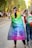 People are walking on a tree-lined street, one person wearing a rainbow flag cape with the word 'Equal' written on it. Others in the background are also casually dressed, potentially attending a public event or parade. The scene conveys an atmosphere of diversity and inclusion.