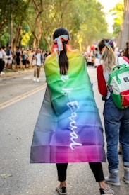 People are walking on a tree-lined street, one person wearing a rainbow flag cape with the word 'Equal' written on it. Others in the background are also casually dressed, potentially attending a public event or parade. The scene conveys an atmosphere of diversity and inclusion.