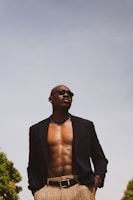 A confident person is standing outdoors, wearing a dark blazer and checkered pants. The blazer is open, revealing a bare chest adorned with a silver necklace and sunglasses. The sky is clear and bright, with trees visible at the edges.