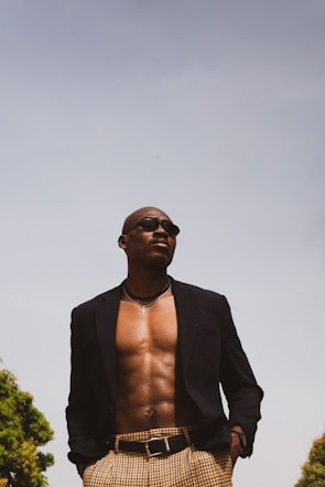 A confident person is standing outdoors, wearing a dark blazer and checkered pants. The blazer is open, revealing a bare chest adorned with a silver necklace and sunglasses. The sky is clear and bright, with trees visible at the edges.