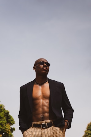A confident person is standing outdoors, wearing a dark blazer and checkered pants. The blazer is open, revealing a bare chest adorned with a silver necklace and sunglasses. The sky is clear and bright, with trees visible at the edges.