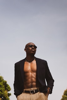 A confident person is standing outdoors, wearing a dark blazer and checkered pants. The blazer is open, revealing a bare chest adorned with a silver necklace and sunglasses. The sky is clear and bright, with trees visible at the edges.
