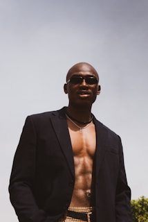A person wearing sunglasses and a black jacket stands confidently with an open chest, showcasing a muscular physique. The background is a clear sky with some greenery visible at the bottom.