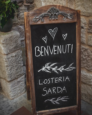 A rustic chalkboard sign with ornate metal and wooden frame stands in front of a stone wall. The sign displays handwritten text in Italian that reads 'Benvenuti' followed by 'L'Osteria Sarda' along with decorative elements such as hearts and leaves.