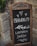 A rustic chalkboard sign with ornate metal and wooden frame stands in front of a stone wall. The sign displays handwritten text in Italian that reads 'Benvenuti' followed by 'L'Osteria Sarda' along with decorative elements such as hearts and leaves.