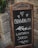 A rustic chalkboard sign with ornate metal and wooden frame stands in front of a stone wall. The sign displays handwritten text in Italian that reads 'Benvenuti' followed by 'L'Osteria Sarda' along with decorative elements such as hearts and leaves.