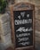 A rustic chalkboard sign with ornate metal and wooden frame stands in front of a stone wall. The sign displays handwritten text in Italian that reads 'Benvenuti' followed by 'L'Osteria Sarda' along with decorative elements such as hearts and leaves.