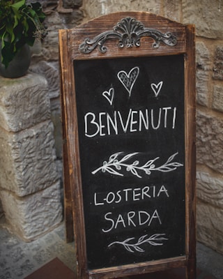 A rustic chalkboard sign with ornate metal and wooden frame stands in front of a stone wall. The sign displays handwritten text in Italian that reads 'Benvenuti' followed by 'L'Osteria Sarda' along with decorative elements such as hearts and leaves.