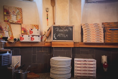A kitchen scene with several stacks of pizza boxes on shelves. There are also plates in a pile, paintings of a chef, and a chalkboard that reads 'Pizzaiolo Attila.' Various kitchen utensils including a rolling pin and a pizza peel are displayed on the wall. The brown and beige color tones give the place a rustic feel.