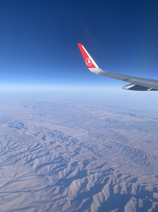 An aircraft wing with a red logo is visible, flying high above a rugged, mountainous desert landscape. The clear sky is a deep blue, transitioning to lighter shades near the horizon.