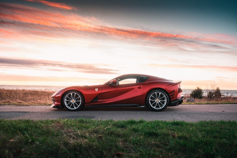 A sleek red sports car is parked on a paved road near the coastline, under a picturesque sunset sky with pink and orange hues. The background features grassy fields and a calm body of water.