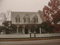 A Victorian-style house stands surrounded by trees and enclosed by a white picket fence. The atmosphere is foggy, creating a muted and vintage ambiance. The foliage on the right tree is a deep red, suggesting autumn.