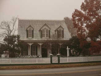A Victorian-style house stands surrounded by trees and enclosed by a white picket fence. The atmosphere is foggy, creating a muted and vintage ambiance. The foliage on the right tree is a deep red, suggesting autumn.