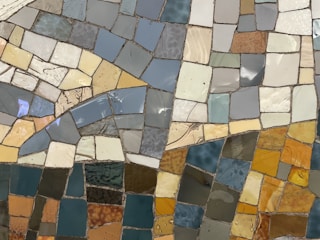 A vibrant mosaic composed of irregularly shaped tiles in various pastel and earthy tones, creating an abstract pattern. The tiles are primarily in shades of blue, yellow, orange, and brown, with a glossy finish that reflects light.
