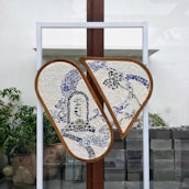 A mosaic artwork mounted on a stand, featuring two semi-circular panels with a blue and white design. The mosaics depict abstract patterns and possibly some calligraphic elements. It is displayed outdoors with potted plants and a stone wall in the background.