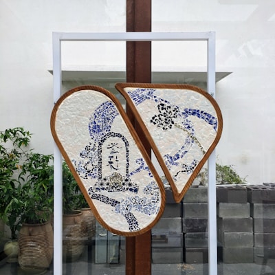 A mosaic artwork mounted on a stand, featuring two semi-circular panels with a blue and white design. The mosaics depict abstract patterns and possibly some calligraphic elements. It is displayed outdoors with potted plants and a stone wall in the background.