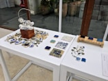 An arrangement of artistic objects on white tables, featuring a central wooden block with a delicate wire sculpture surrounded by broken ceramic pieces. There are several tile samples in various shades, alongside scattered ceramic fragments. The setting appears to be a modern indoor space, with large glass windows and potted plants visible in the background.