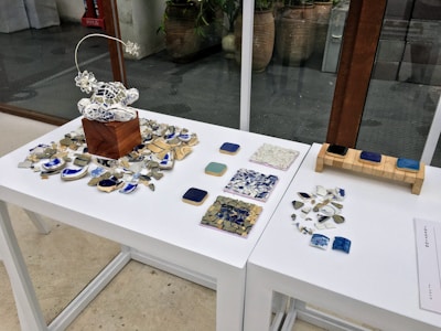 An arrangement of artistic objects on white tables, featuring a central wooden block with a delicate wire sculpture surrounded by broken ceramic pieces. There are several tile samples in various shades, alongside scattered ceramic fragments. The setting appears to be a modern indoor space, with large glass windows and potted plants visible in the background.